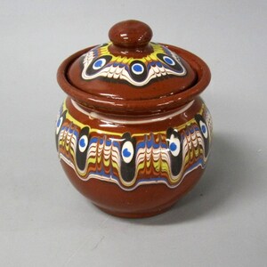 May include: A brown ceramic jar with a matching lid, featuring a decorative marbled pattern in yellow, blue, white, and black around the body and lid. The jar has a rounded shape with a small knob handle on the center of the lid.