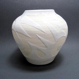 May include: A cream-colored ceramic vase with a rounded body and a wide opening. The vase features a raised, white, flame-like pattern around its exterior. The design is symmetrical and adds a decorative element to the vessel.