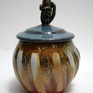 May include: A ceramic jar with a blue lid and a decorative bronze handle. The jar's body features vertical stripes of brown, white, and tan. The jar is a decorative piece.