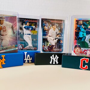 May include: Baseball trading cards featuring Francisco Lindor, Shohei Ohtani, Aaron Judge, and Steven Kwan. The cards are displayed in protective cases, resting on blue, black, and green team logo holders. The Mets, Dodgers, Yankees, and Guardians logos are visible.
