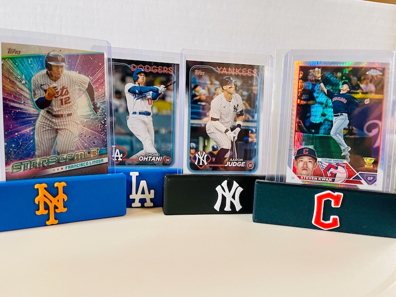 May include: Baseball trading cards featuring Francisco Lindor, Shohei Ohtani, Aaron Judge, and Steven Kwan. The cards are displayed in protective cases, resting on blue, black, and green team logo holders. The Mets, Dodgers, Yankees, and Guardians logos are visible.