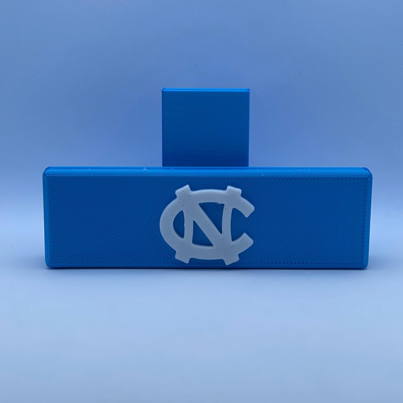 May include: A light blue rectangular box with a white emblem, and a smaller blue square behind it. The emblem is a stylized white letter "N" and "C" intertwined. The box is made of a textured material.