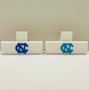 May include: Two white rectangular objects with a blue and a light blue logo. Each object has a small white square on top. The logos are the same design, a stylized letter "N" and "C" intertwined. The objects are on a white surface.