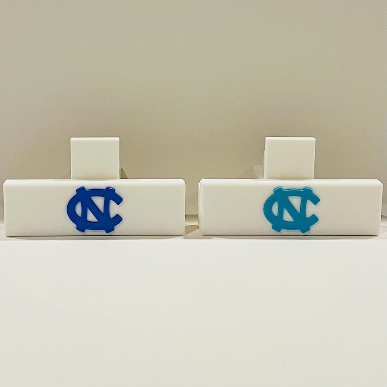 May include: Two white rectangular objects with a blue and a light blue logo. Each object has a small white square on top. The logos are the same design, a stylized letter "N" and "C" intertwined. The objects are on a white surface.