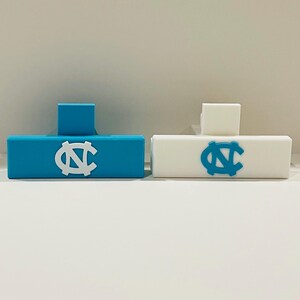 May include: Two rectangular objects, one light blue and one white, each with a raised square and a logo. The light blue object has a white logo, and the white object has a light blue logo. The logo is a stylized letter "N" and "C".