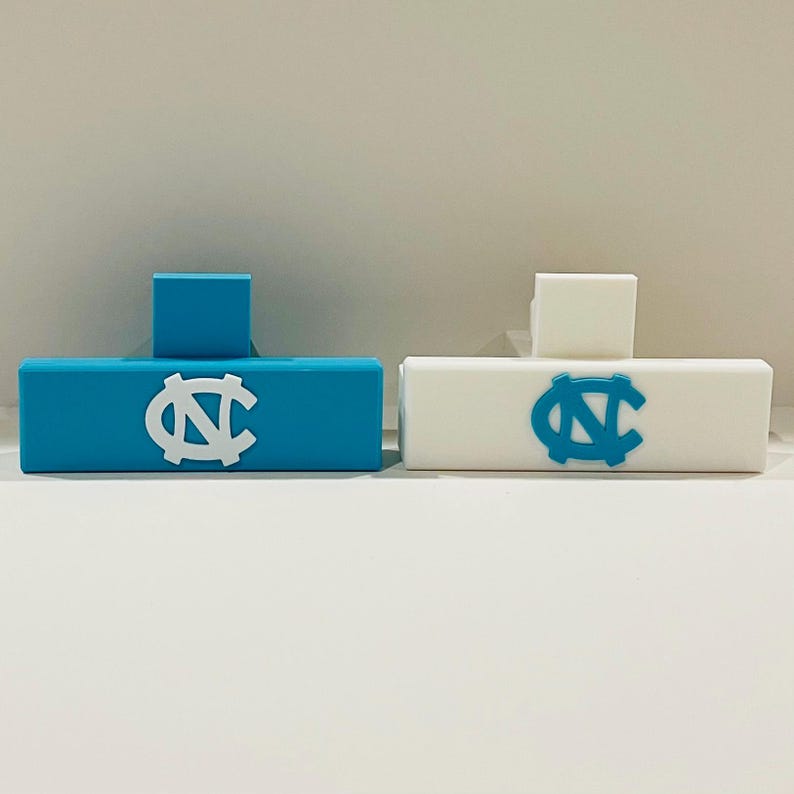 May include: Two rectangular objects, one light blue and one white, each with a raised square and a logo. The light blue object has a white logo, and the white object has a light blue logo. The logo is a stylized letter "N" and "C".