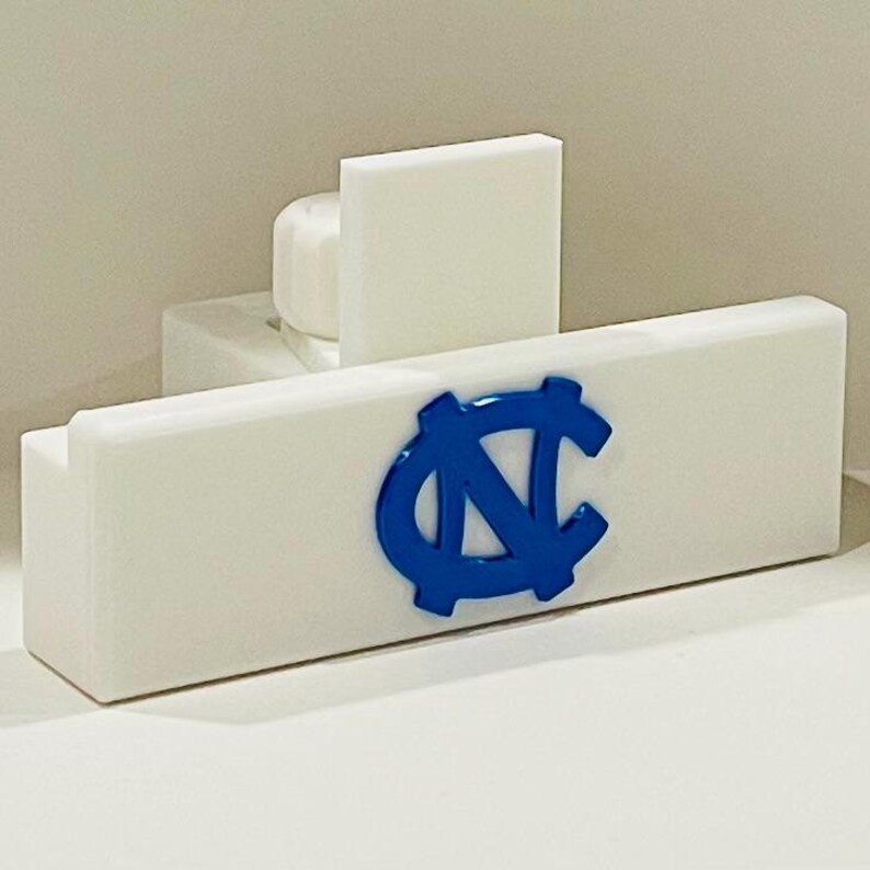 May include: A white rectangular object with a blue logo, possibly a door stop or decorative item. The logo is a stylized blue "NC" with a gear-like border. The object has a cylindrical piece and a rectangular piece attached to it.