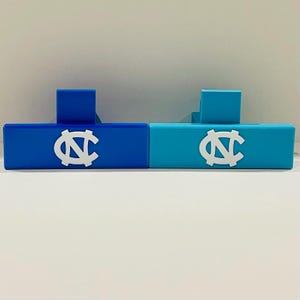 May include: Two rectangular, blue and teal-colored decorative blocks. Each block features a white logo. The blocks have a small, square support on the back. The blocks are displayed on a white surface.
