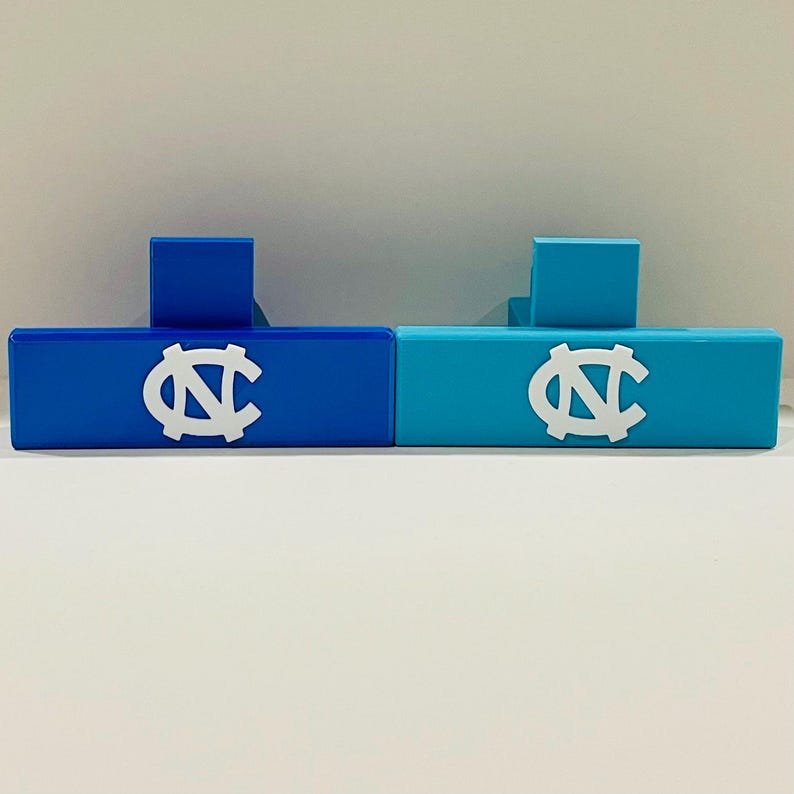 May include: Two rectangular, blue and teal-colored decorative blocks. Each block features a white logo. The blocks have a small, square support on the back. The blocks are displayed on a white surface.