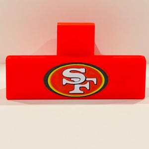 Peut inclure: Un objet rouge vif en forme de T, avec une base rectangulaire et une partie verticale. La base présente un emblème ovale avec les lettres "SF" blanches, bordées d'or et de noir. Il s'agit probablement d'un article sportif.