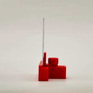 May include: A red plastic tool with a vertical metal blade. The tool has a rectangular base with a cylindrical knob and a slot. The blade is thin and silver, extending upwards from the base.
