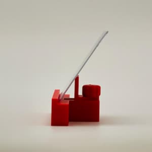 May include: A bright red phone stand with a rectangular base and a vertical support holding a clear, rectangular screen. The stand is designed to hold a phone or tablet at an angle for easy viewing. The stand is made of plastic.