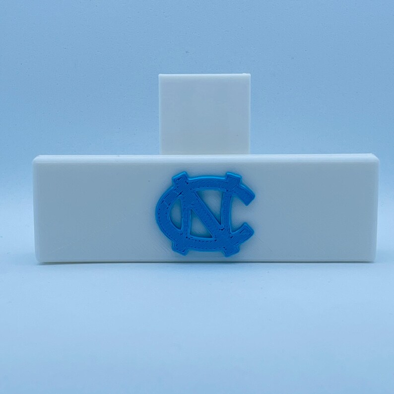 May include: A white rectangular object with a light blue logo of a stylized letter "N" and "C" intertwined. A smaller white rectangular object is behind the larger one. The logo is in the center of the larger object.