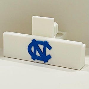 May include: A white rectangular object with a blue logo featuring the letters "N" and "C" intertwined. The object has a rectangular base and a smaller, upright rectangular piece attached to the back.