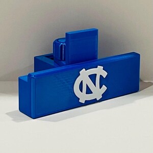 May include: A blue and white decorative item featuring the University of North Carolina logo. The item is a rectangular shape with a raised logo and a vertical piece attached to the top. The item is made of a solid material.