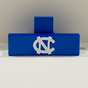 May include: A blue rectangular object with a white logo, featuring a stylized "N" and "C" intertwined. A smaller blue rectangular piece is attached to the top. The object is likely a decorative item or a holder.
