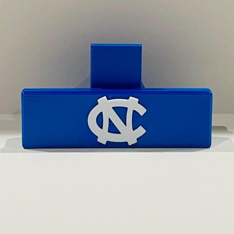 May include: A blue rectangular object with a white logo, featuring a stylized "N" and "C" intertwined. A smaller blue rectangular piece is attached to the top. The object is likely a decorative item or a holder.