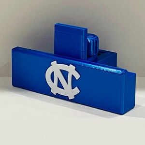 May include: A blue rectangular object with a white logo, possibly a sports-related item. The logo is a stylized letter "N" and "C" intertwined. The object has a boxy design and is made of a solid, possibly plastic, material.