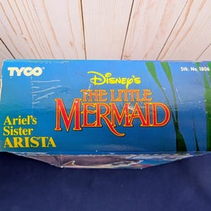 Może przedstawiać: Niebieskie pudełko z napisem "Disney's The Little Mermaid" w kolorze czerwonym i ż&oacute;łtym. Pudełko zawiera r&oacute;wnież napis "Ariel's Sister Arista" i logo Tyco. Tło przedstawia podwodną scenerię.