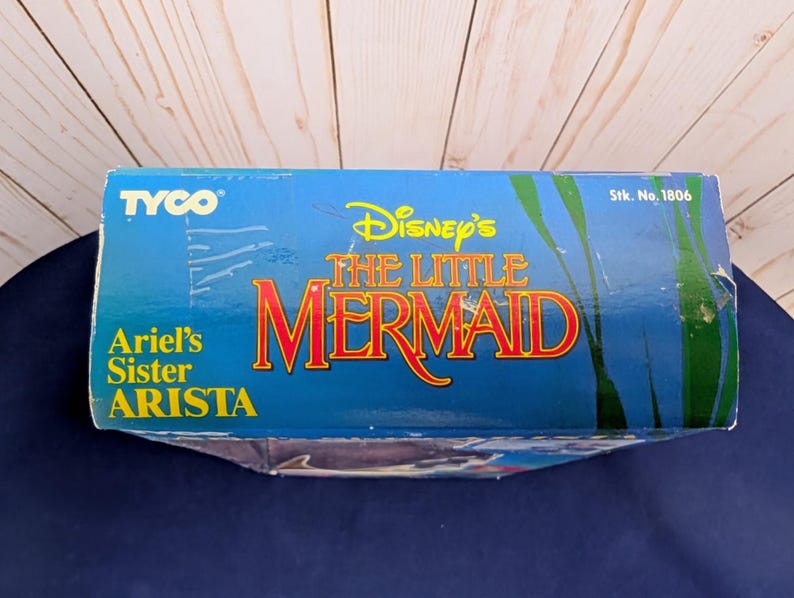 Może przedstawiać: Niebieskie pudełko z napisem "Disney's The Little Mermaid" w kolorze czerwonym i ż&oacute;łtym. Pudełko zawiera r&oacute;wnież napis "Ariel's Sister Arista" i logo Tyco. Tło przedstawia podwodną scenerię.