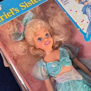 Może przedstawiać: Lalka siostry Arielki, Aristy, w pudełku. Lalka ma blond włosy i ubrana jest w niebieski str&oacute;j z kokardą. Na pudełku widnieje napis "Ariel's Sister Arista". Lalka jest ubrana na Podwodną Imprezę.