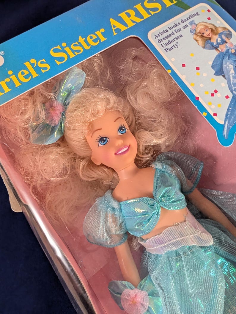 Może przedstawiać: Lalka siostry Arielki, Aristy, w pudełku. Lalka ma blond włosy i ubrana jest w niebieski str&oacute;j z kokardą. Na pudełku widnieje napis "Ariel's Sister Arista". Lalka jest ubrana na Podwodną Imprezę.