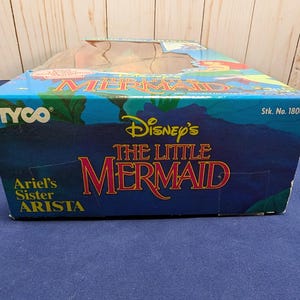 Może przedstawiać: Niebieskie pudełko z logo Disney's The Little Mermaid w kolorze czerwonym i złotym. Pudełko ma r&oacute;wnież logo Tyco i napis "Ariel's Sister Arista". Pudełko znajduje się na niebieskiej powierzchni.