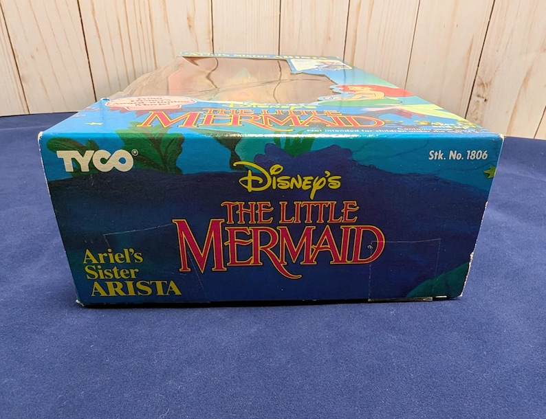 Może przedstawiać: Niebieskie pudełko z logo Disney's The Little Mermaid w kolorze czerwonym i złotym. Pudełko ma r&oacute;wnież logo Tyco i napis "Ariel's Sister Arista". Pudełko znajduje się na niebieskiej powierzchni.