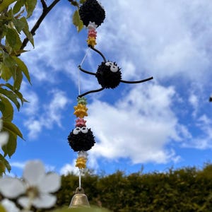 Puede incluir: Un carillón negro y blanco con una campana en la parte inferior. El carillón presenta tres bolas negras y peludas con ojos blancos y una cadena de cuentas de colores.