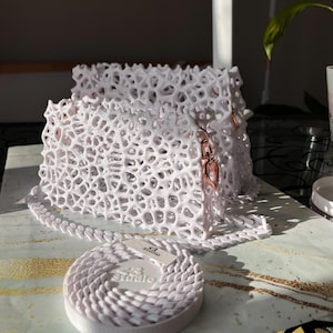 May include: A white, lacy,  shoulder bag with a rose gold chain strap and hardware. The bag is made of a 3D printed material and has a unique, organic design. The bag is sitting on a white surface with a white, coiled chain strap.