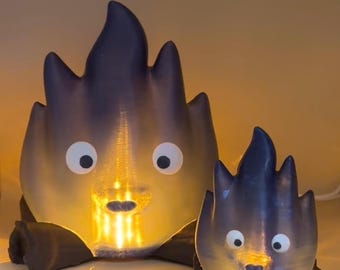 May include: Two glowing, 3D printed, campfire figurines with googly eyes and a black base. The larger figurine is in the background and the smaller figurine is in the foreground.