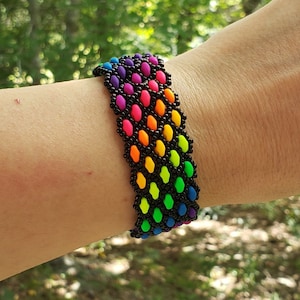 Op de afbeelding: Een regenboogkleurige kralen armband met zwarte kralen. De armband zit om een pols. De achtergrond is wazig en toont groen gebladerte.