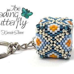 Peut inclure: Un porte-clés en forme de cube bleu, blanc et orange avec un anneau et une chaîne argentés. Le cube présente un motif géométrique et est attaché à un anneau et une chaîne argentés. Le porte-clés est étiqueté "The Beading Butterfly © Kassie Shaw".