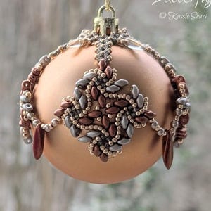 Puede incluir: Un adorno navideño artesanal con una base de color melocotón, adornado con intrincados abalorios en tonos marrón, plateado y dorado. El adorno está suspendido por un clip dorado. El texto "The Beading Butterfly" es visible.