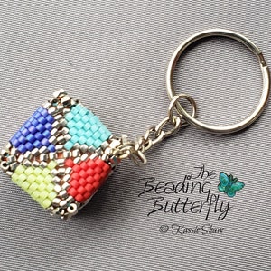May include: A beaded keyring with a geometric design. The square charm has sections of blue, turquoise, red, and yellow beads, with silver metal accents and a keyring. The text "The Beading Butterfly" is visible.