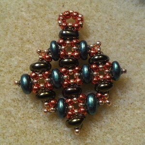 K&ouml;nnte beinhalten: Ein kleiner, filigraner Anh&auml;nger aus Kupfer-, Gold- und blaugr&uuml;nen Perlen. Der Anh&auml;nger hat die Form eines Sterns mit einer &Ouml;se oben zum Aufh&auml;ngen.