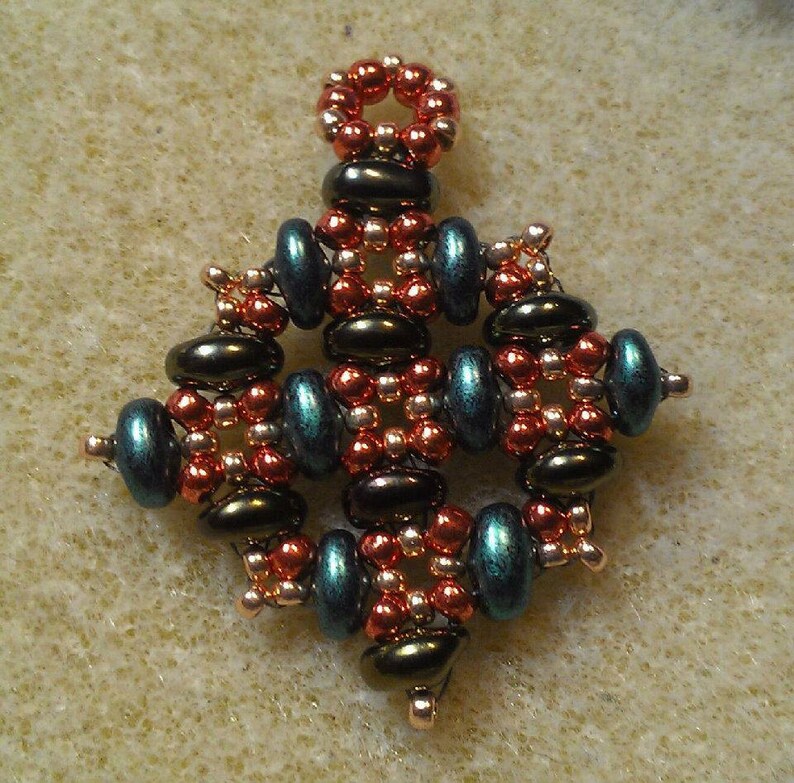 K&ouml;nnte beinhalten: Ein kleiner, filigraner Anh&auml;nger aus Kupfer-, Gold- und blaugr&uuml;nen Perlen. Der Anh&auml;nger hat die Form eines Sterns mit einer &Ouml;se oben zum Aufh&auml;ngen.