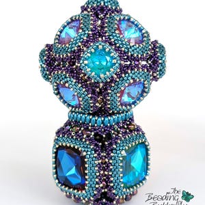 May include: A handcrafted beaded bottle stopper in shades of teal, purple, and blue. The stopper features large, faceted blue crystals and intricate beadwork. The design includes a rounded top and a cylindrical base, showcasing a detailed, artistic creation.