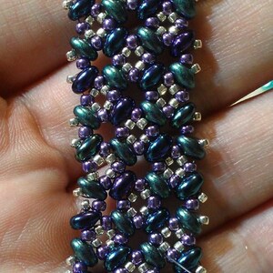 K&ouml;nnte beinhalten: Nahaufnahme eines Armbands mit Perlen in einem geometrischen Muster. Die Perlen sind in Gr&uuml;n-, Lila- und Silbert&ouml;nen.