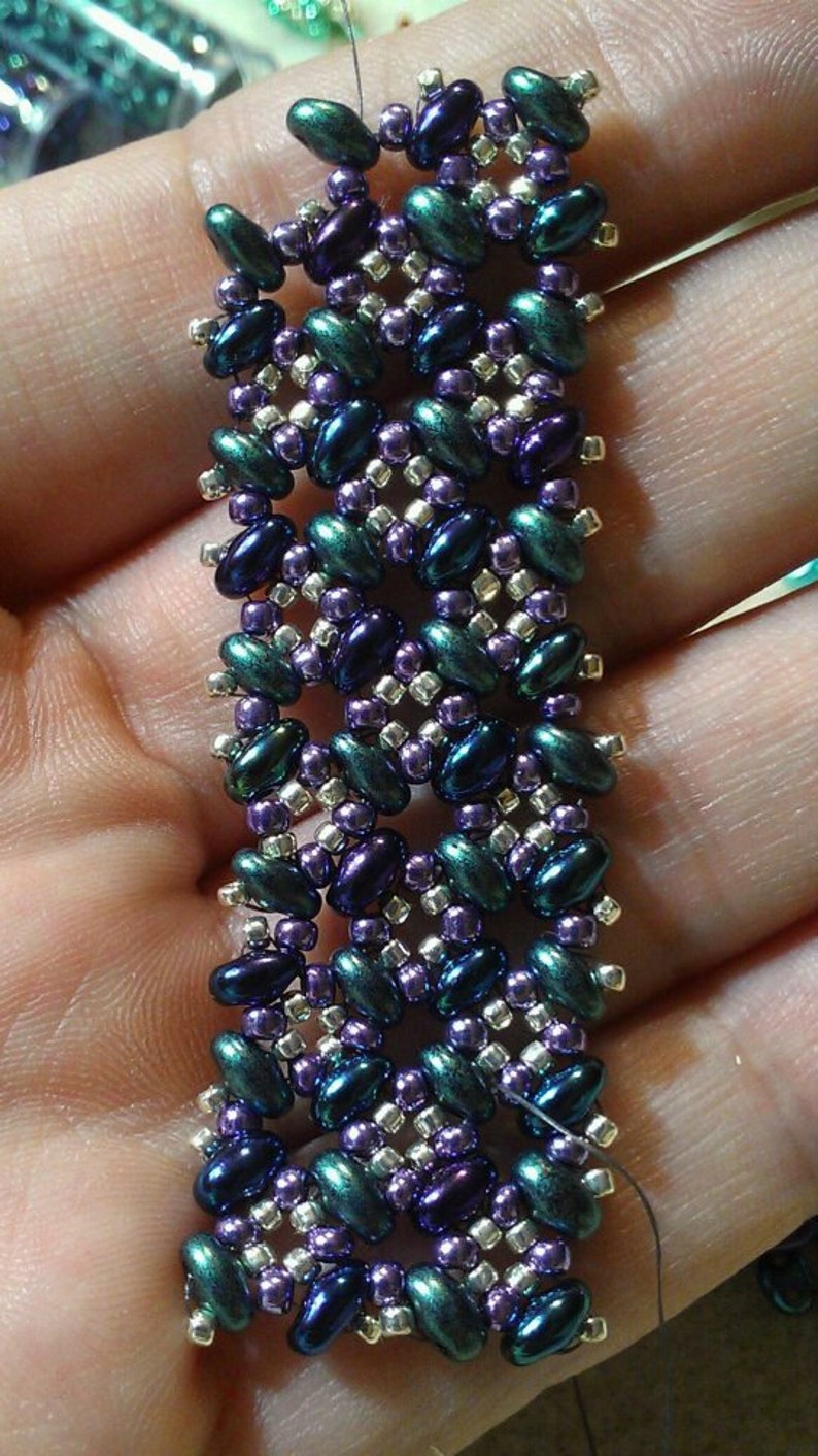 K&ouml;nnte beinhalten: Nahaufnahme eines Armbands mit Perlen in einem geometrischen Muster. Die Perlen sind in Gr&uuml;n-, Lila- und Silbert&ouml;nen.