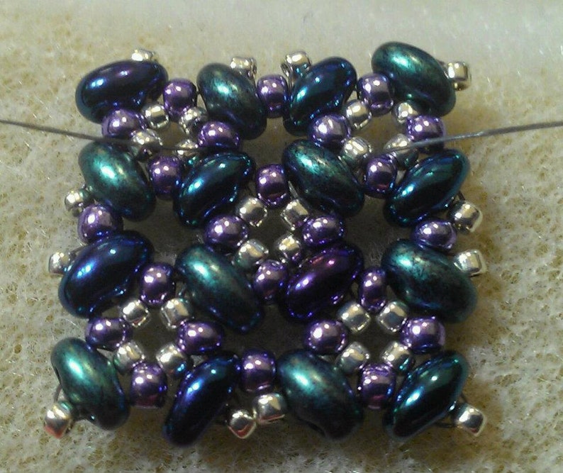 K&ouml;nnte beinhalten: Ein quadratischer Perlenanh&auml;nger mit einem sich wiederholenden Muster aus dunkelblauen, violetten und silbernen Perlen.