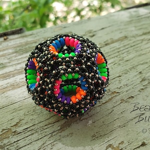 May include: A spherical beaded object with a black base, accented with colourful rings of blue, green, orange, pink, and purple. The object is covered in small silver beads. The Beading Butterfly logo is visible.