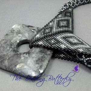 Puede incluir: Un collar de cuentas negras y plateadas con un gran colgante cuadrado gris. El colgante tiene un agujero en el centro. El collar está anudado en la parte delantera y los extremos están atados juntos.