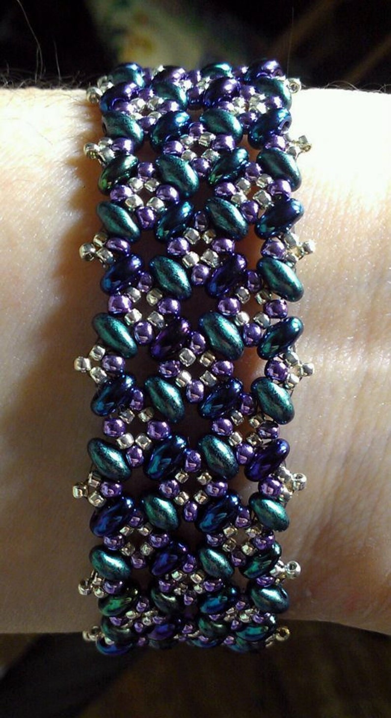 K&ouml;nnte beinhalten: Ein Armband mit Perlen in einem geometrischen Muster in den Farben Lila, T&uuml;rkis und Silber.