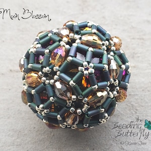 Könnte beinhalten: Eine Perlenkugel mit geometrischem Design, bestehend aus einer Mischung aus dunkelgrünen, silbernen, goldenen und violetten Perlen. Die Kugel heißt "Moon Blossom" und wurde von "The Beading Butterfly" hergestellt.