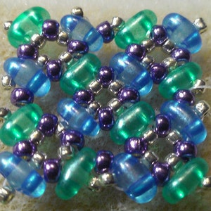 K&ouml;nnte beinhalten: Ein quadratischer Perlenanh&auml;nger mit einem sich wiederholenden Muster aus blauen, gr&uuml;nen und violetten Perlen. Die Perlen sind in einem symmetrischen Design angeordnet.