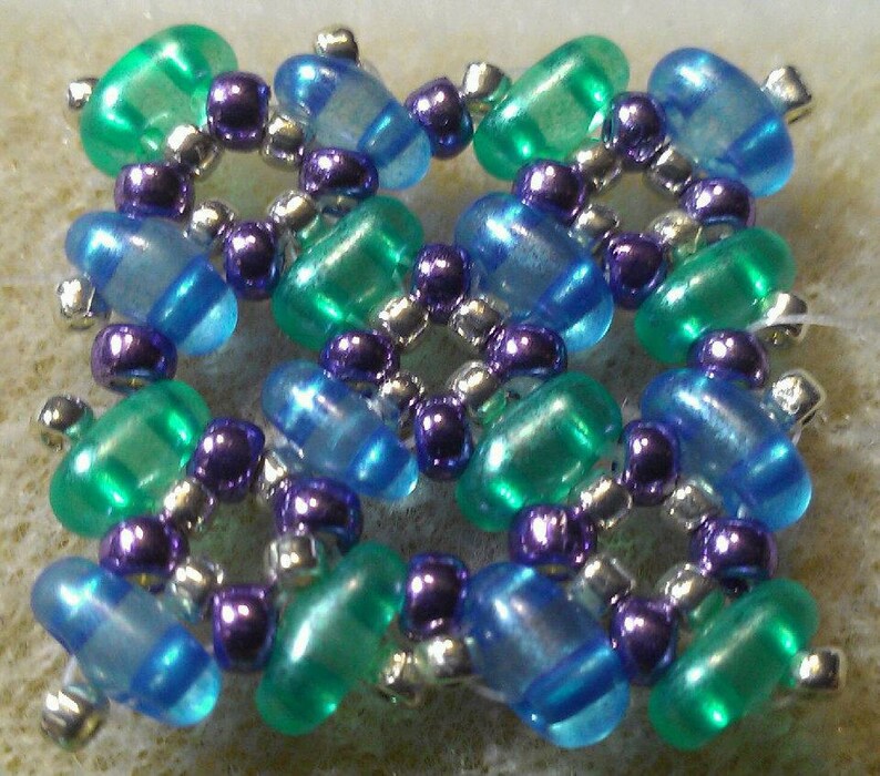 K&ouml;nnte beinhalten: Ein quadratischer Perlenanh&auml;nger mit einem sich wiederholenden Muster aus blauen, gr&uuml;nen und violetten Perlen. Die Perlen sind in einem symmetrischen Design angeordnet.