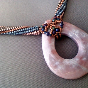 May include: A necklace with a pink and white stone pendant. The pendant is shaped like a circle with a hole in the middle. The necklace is made of small blue, copper, and iridescent beads.