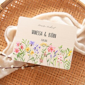 May include: A wooden wedding announcement with the names "VANESSA & BJ&Ouml;RN" and the date "25.05.2026" in black text. The announcement features a floral border with pink, yellow, and purple flowers. The text "Unsere Hochzeit" is above the names.