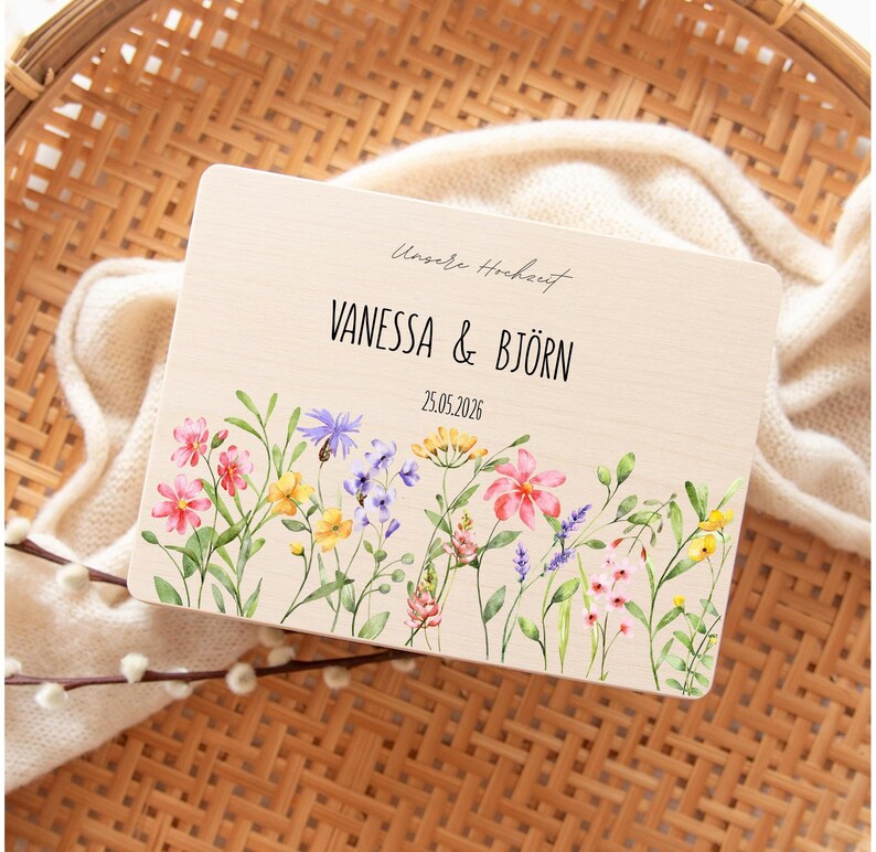 May include: A wooden wedding announcement with the names "VANESSA & BJ&Ouml;RN" and the date "25.05.2026" in black text. The announcement features a floral border with pink, yellow, and purple flowers. The text "Unsere Hochzeit" is above the names.